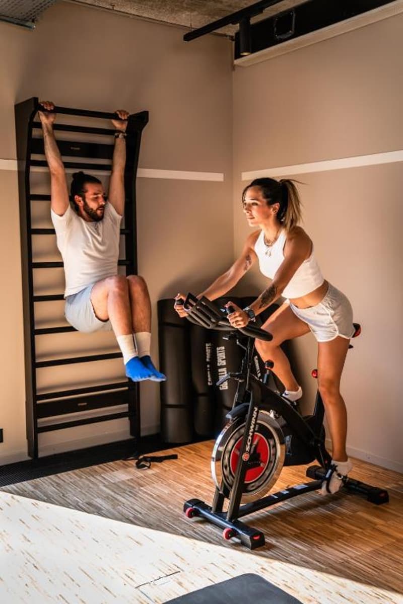 The image shows a man doing pull-ups on a wall-mounted exercise bar, and a woman riding an exercise bike in a home gym setting. The room has beige walls and a wooden floor.