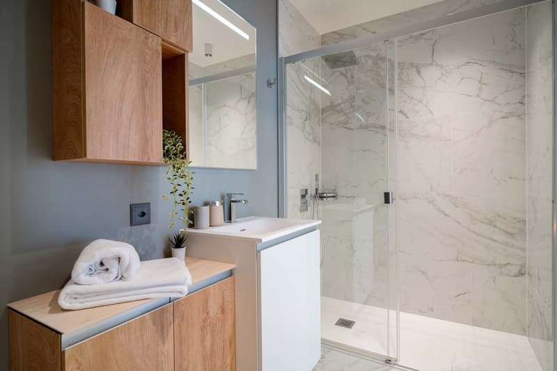 Modern bathroom with marble-like tile walls, a glass shower enclosure, a vanity with a sink, and wooden storage cabinets. There are towels and a potted plant visible as well.