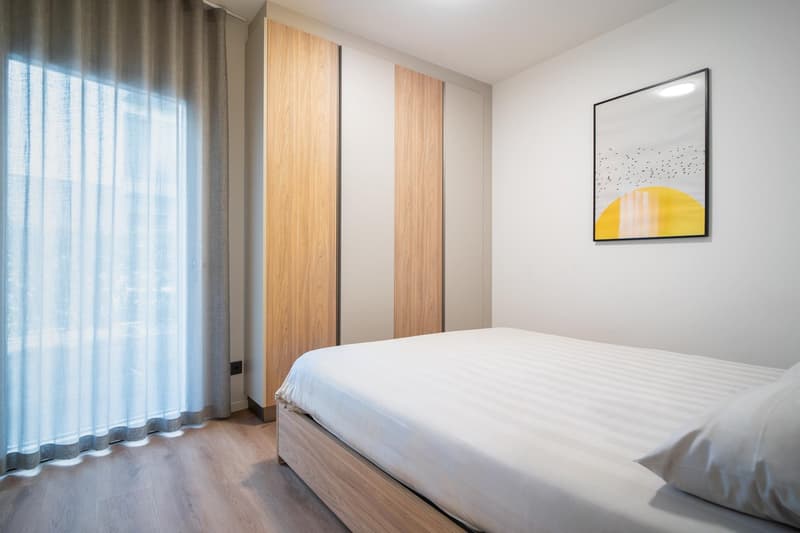 Single bed with white sheets, a large window with white curtains, a wall-mounted painting, wooden wardrobe