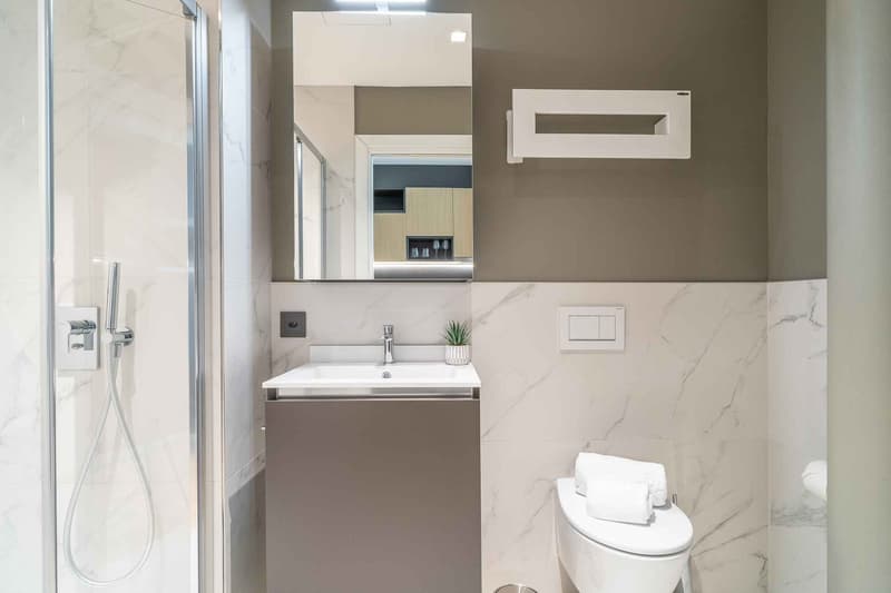 Modern bathroom with a walk-in shower, a white sink with a faucet, a mirror, and a toilet. The floor and walls are covered with white marble tiles.