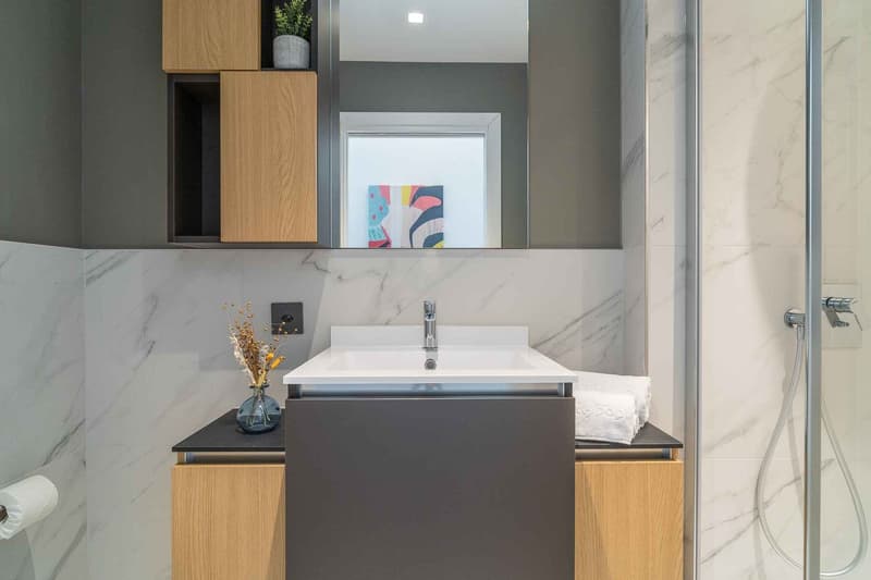 modern bathroom with marble tiles, white sink, wooden cabinets, shower enclosure, mirror, power outlet, toilet paper holder