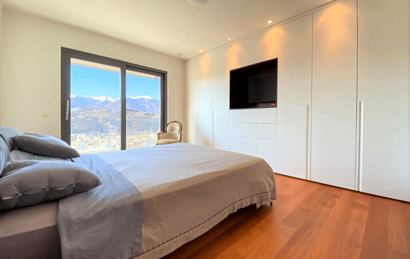 Spacious bedroom with large windows offering a scenic view of the city, hardwood flooring, white walls, built-in closet, and a mounted TV on the wall.
