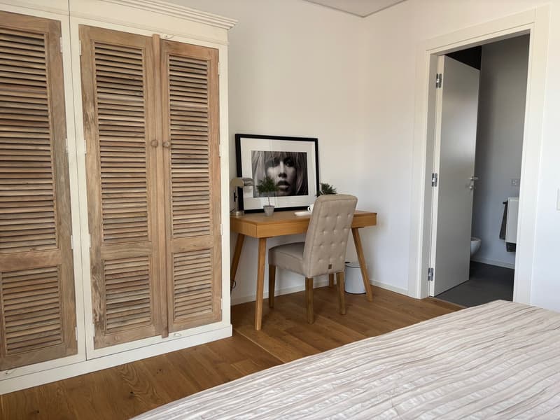 Spacious bedroom with wooden louvered closet doors, a wooden desk with a framed artwork, and a tufted upholstered chair. The room has hardwood flooring and a neutral color scheme.