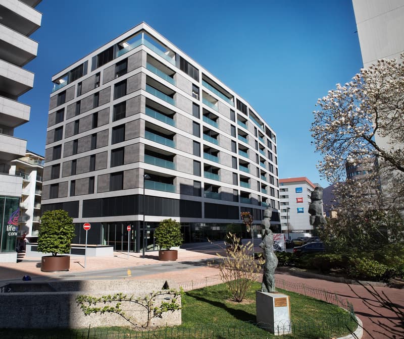 Modern multi-story building with glass and concrete facade, balconies, and a statue in the foreground. The building appears to be a commercial or residential property, surrounded by landscaping and other buildings.