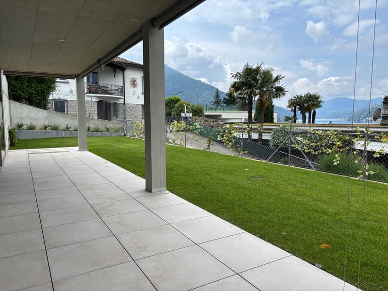 Spacious outdoor patio with tiled flooring, overlooking a lush garden with palm trees and a scenic mountain view in the background. The property appears to be a multi-story building with a balcony or terrace visible.