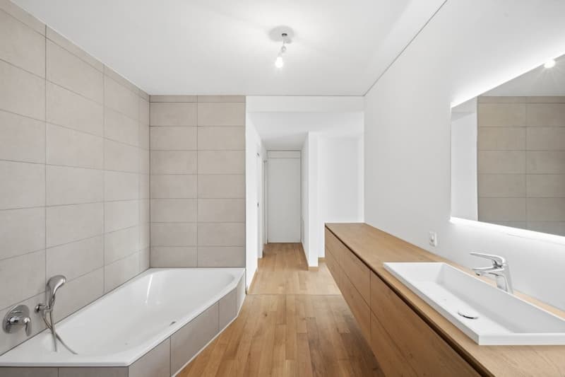 Modern minimalist bathroom with wooden floor, white bathtub, modern vanity, large mirror, and silver fixtures.