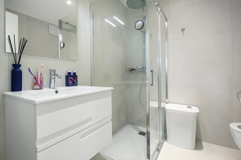 Modern bathroom with white cabinets, glass shower, floor-level WC, white tiles, modern sink with faucet, toiletries, mirror above sink, waste basket, trash can