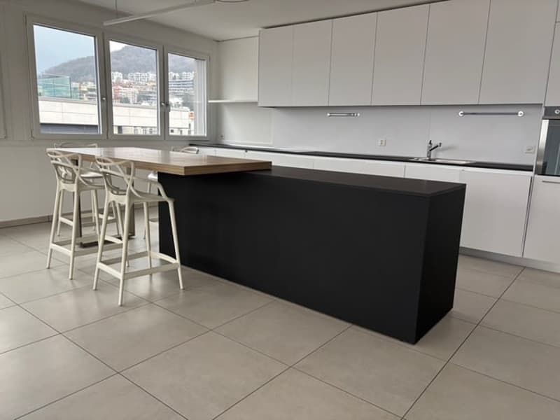 Modern kitchen, black island, white tiles, large windows, wooden bar counter, silver faucet, three white stools.