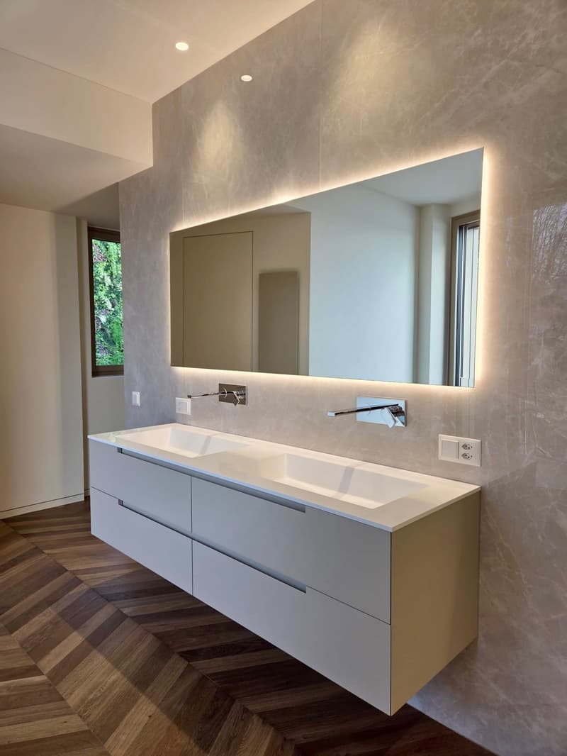 A modern bathroom with marble wall and wooden floor. A large mirror is mounted on the wall. Two sinks with double faucets are placed on top of a white cabinet. There are two outlets on the wall