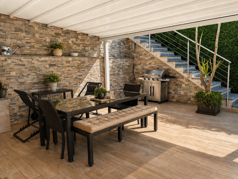 Outdoor dining area with a glass table, cushioned bench, and chairs. The area is covered by a pergola with brick walls and a staircase. There is a gas grill and potted plants.