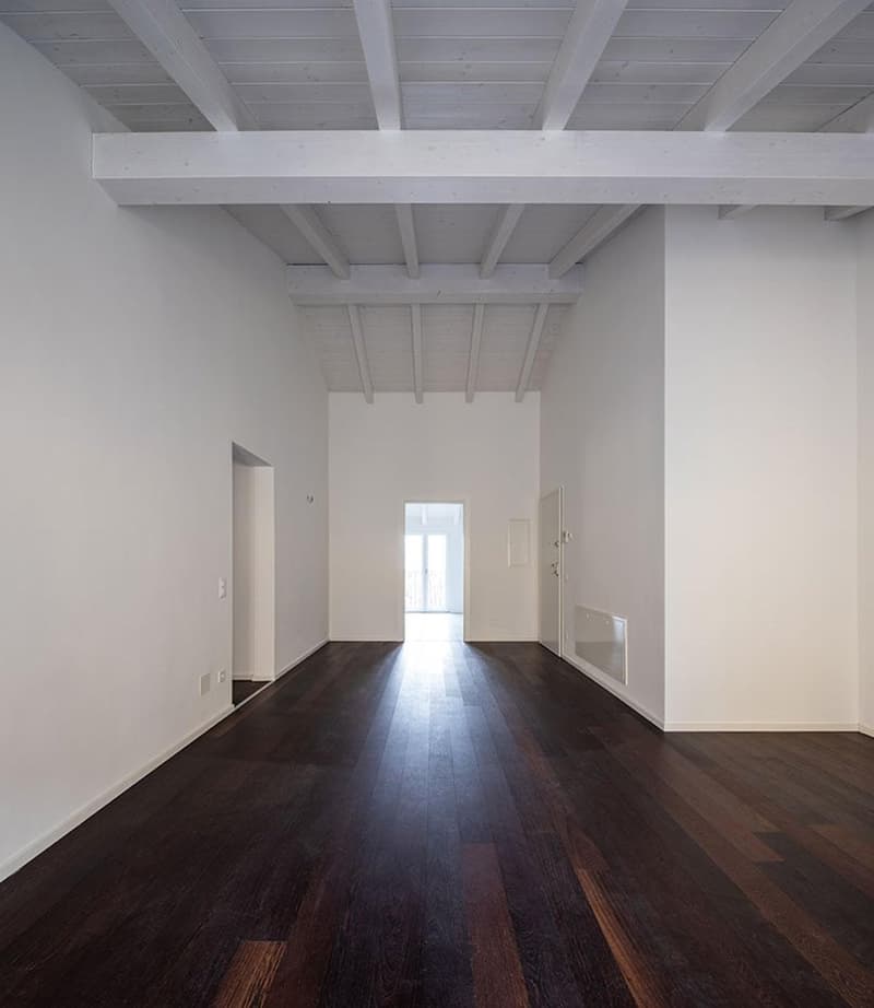 Empty room, white walls, wooden floor, white ceiling beams, doors on both sides, light coming from the end