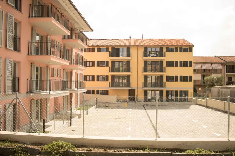 Two buildings, one pink and one yellow, both with balconies, multiple windows, and a fenced parking area in front.