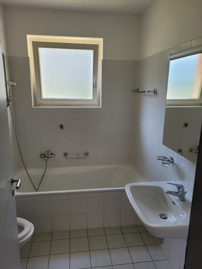 The image shows a bathroom with a bathtub, sink, and toilet. The room has white tile walls and floors, and a window with frosted glass. The bathroom appears to be clean and well-maintained.