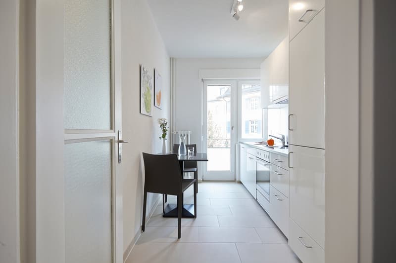 Bright and modern kitchen with white cabinets, black countertop, and a dining table. The kitchen has a dishwasher and large windows providing natural light.