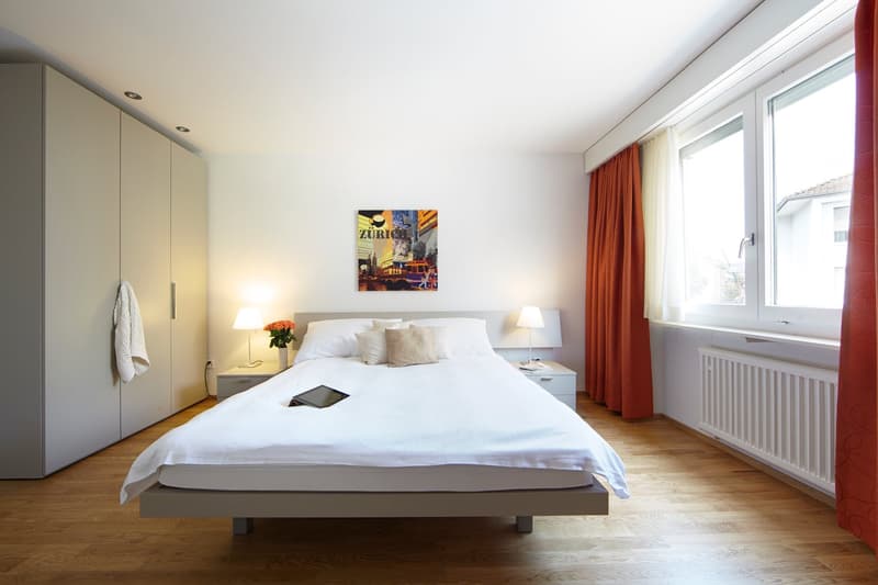 Spacious bedroom with a large bed, white bedding, and a modern artwork on the wall. The room has large windows with red curtains, providing natural light. There is a built-in wardrobe along one wall and a bedside table with lamps on either side of the bed