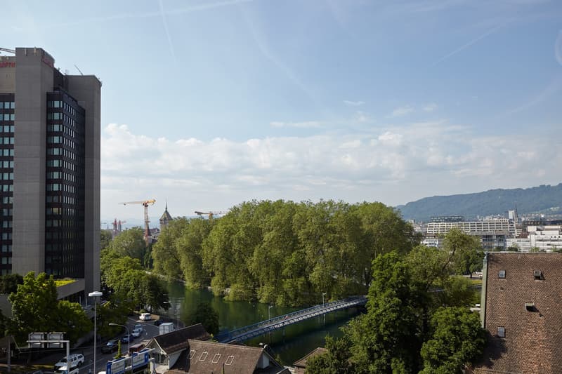 The image shows a cityscape with a river running through it. There are tall buildings, including a high-rise office building, as well as trees and other structures visible. The sky is partly cloudy, and the overall scene appears to be a view of an urban a