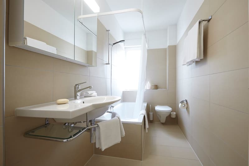 Spacious bathroom with white tiles, large vanity sink, mirror cabinet, shower, and toilet. Towels are provided.