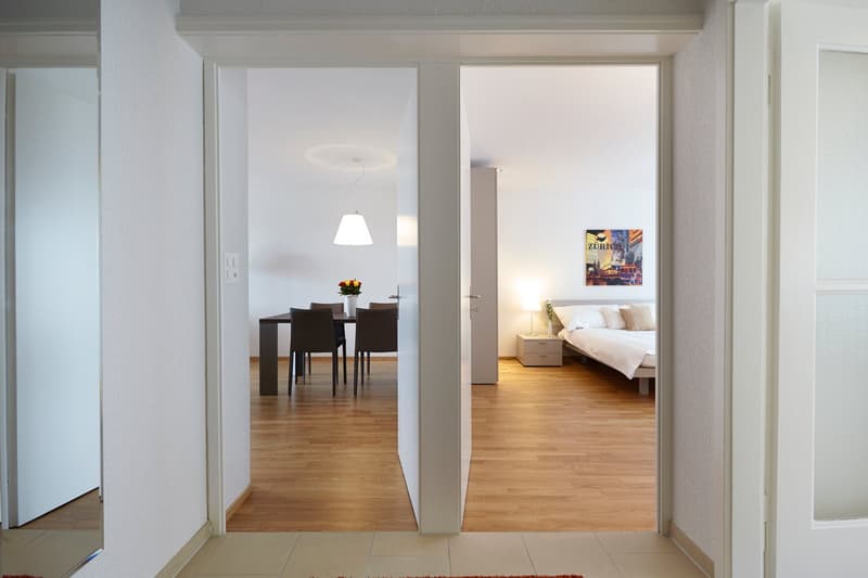 Open floor plan with living room, dining room, and bedroom visible. Hardwood floors throughout, white walls, and modern furnishings. Dining table with 4 chairs, pendant light fixture, and artwork on the wall. Bedroom has a bed, nightstand, and artwork on