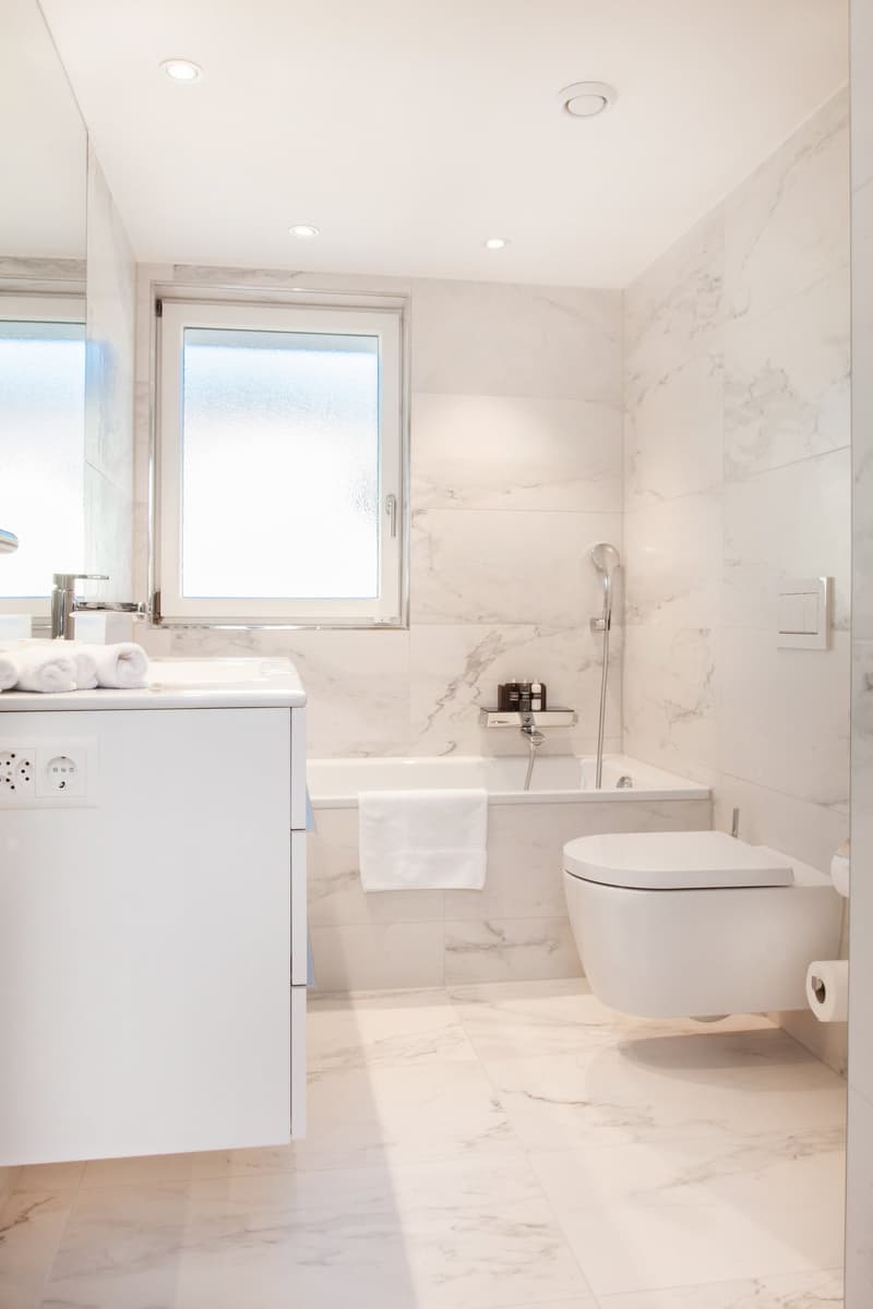 all white bathroom with bathtub, toilet, large mirror, white marble floors