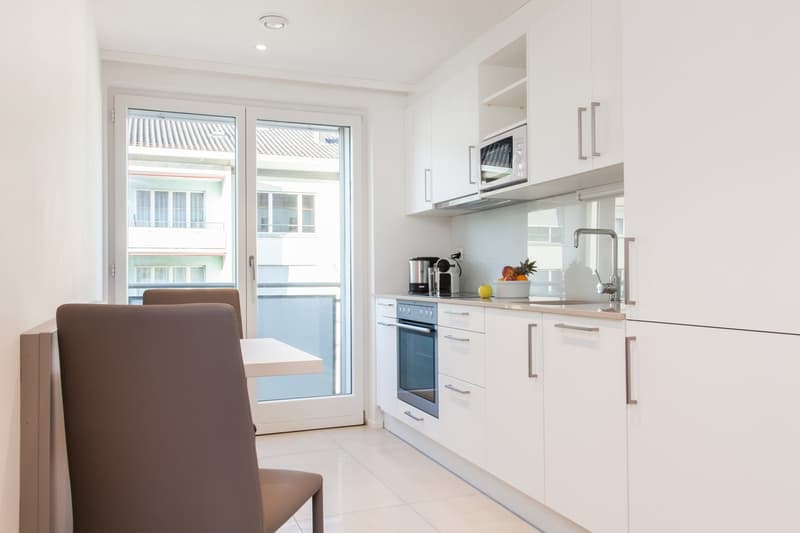 Open-plan kitchen, modern white cabinetry, glass door leading to balcony, granite countertops, integrated appliances, island seating, natural light