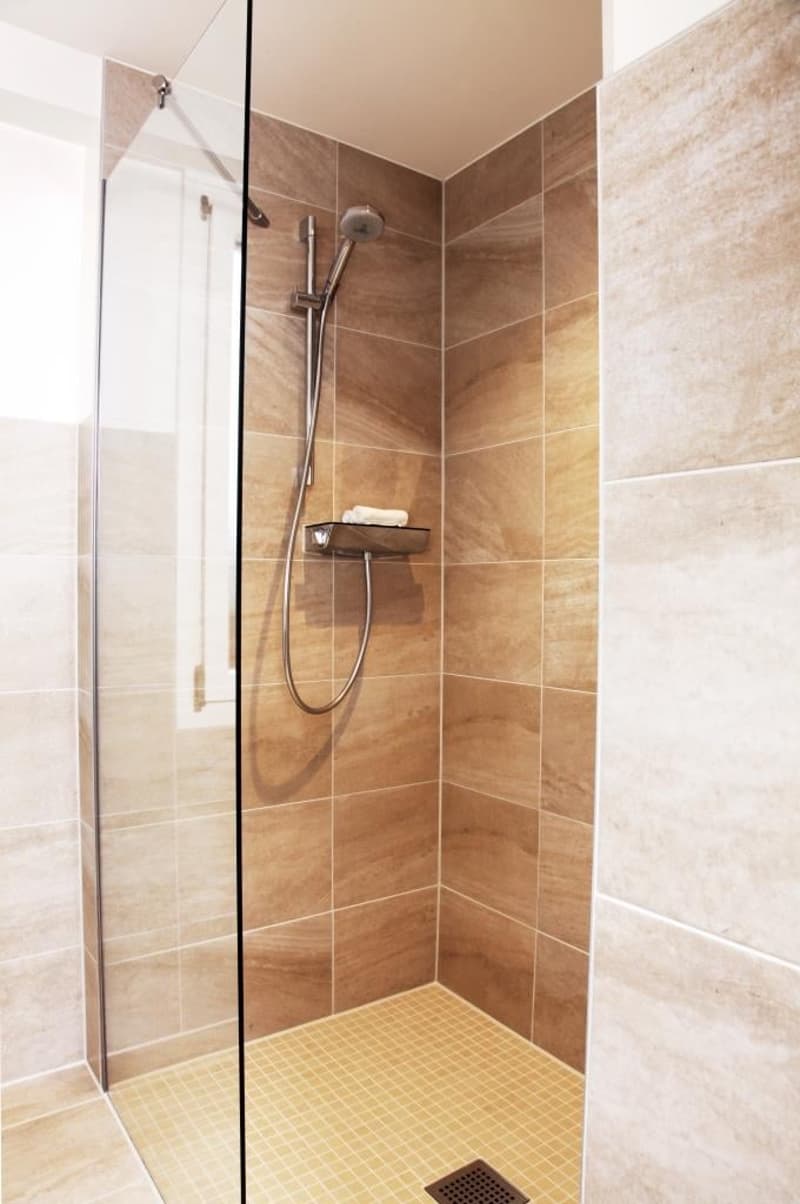 Spacious shower area with glass door, tiled walls in beige and brown tones, shower head and fixtures, towel shelf