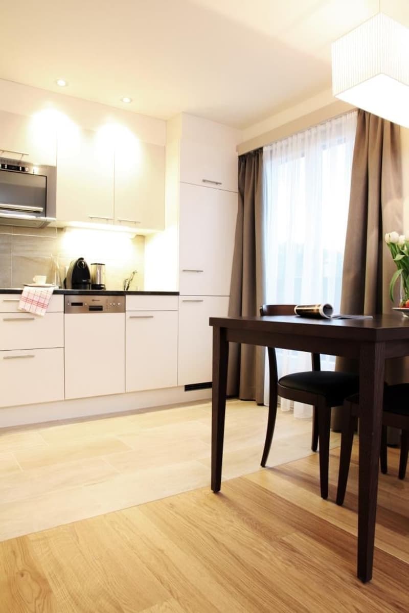The image shows a modern kitchen and dining area. The kitchen features white cabinets, a dishwasher, and other appliances. There is a wooden dining table with chairs in the adjacent dining area. The room has large windows that provide natural light, and t