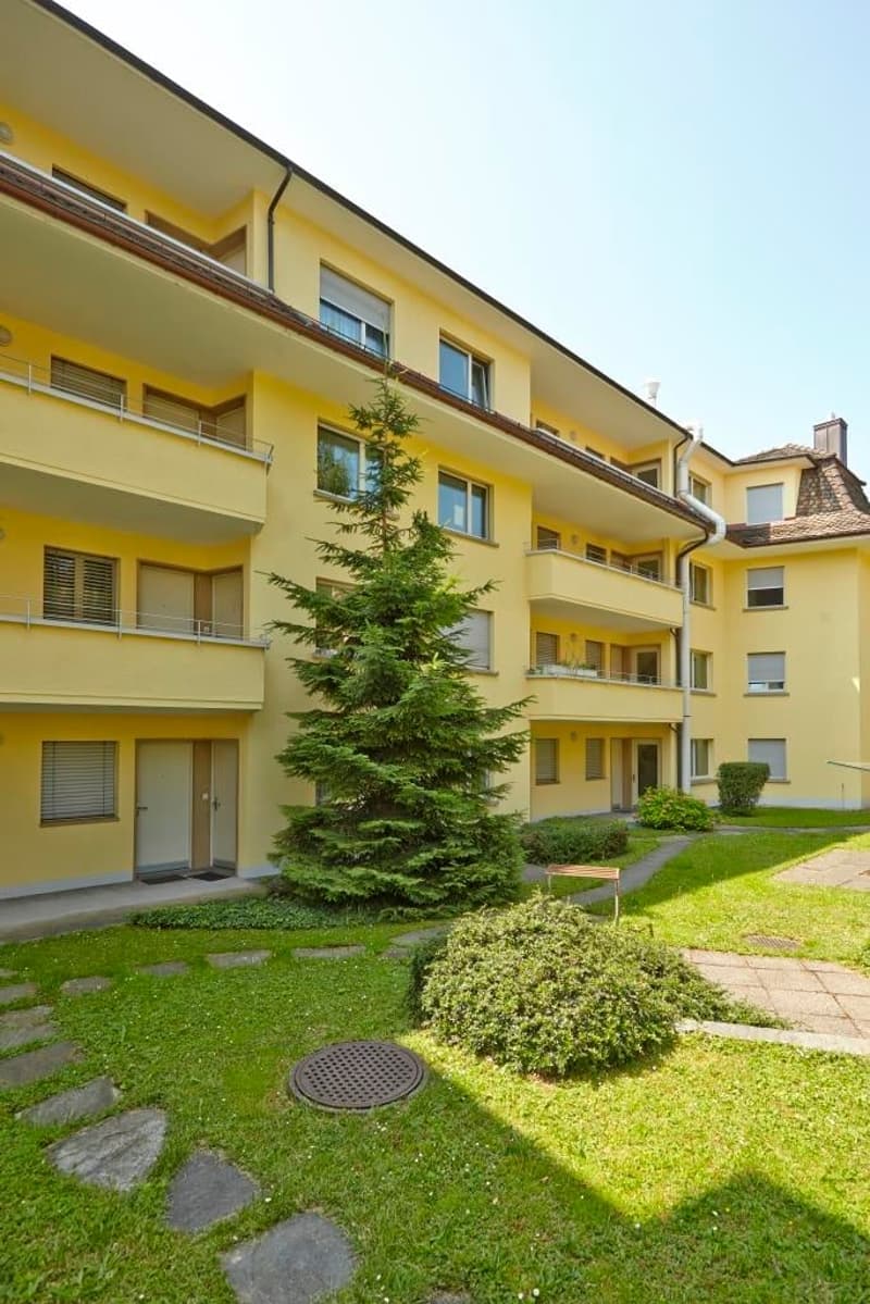 3 story apartment building, yellow exterior, multiple balconies, window shutters, entryway with steps, tree, walkway, manhole cover, grass, bushes
