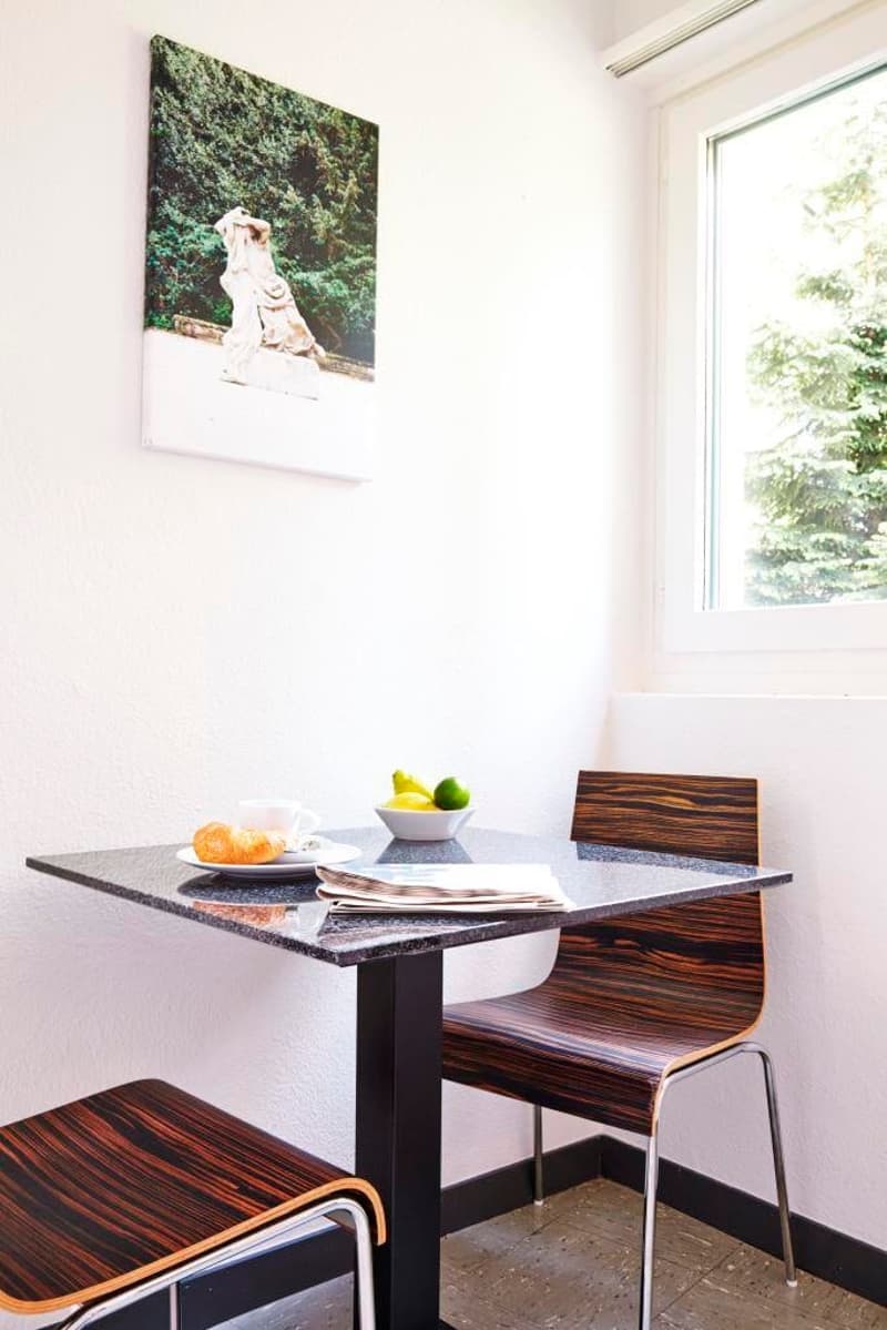 Dining table with chairs, bowl of fruit, a cup, croissant on a plate, magazine on the table, a framed art piece, large window with a view of trees
