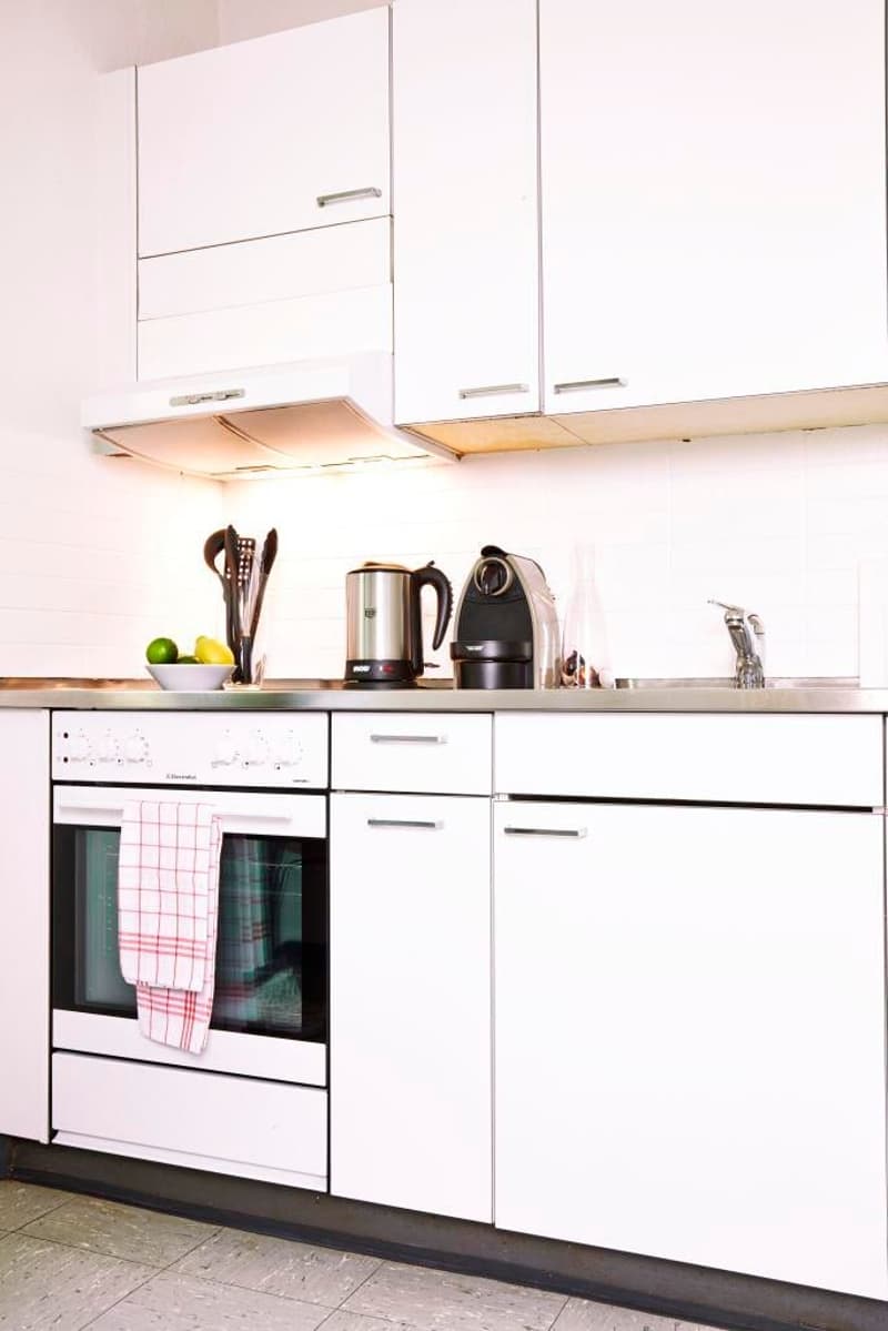Modern white kitchen with built-in appliances, stainless steel countertop, built-in oven, toaster, coffee maker, kettle, bowl of fruits, cutlery set, faucet, sink, tiled flooring, cabinets
