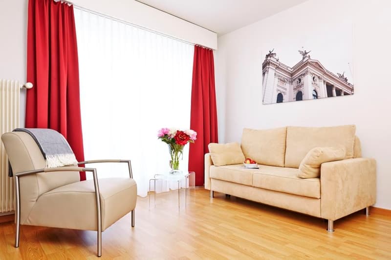 tan leather armchair, small table with flowers, cream couch with pillows, white wall, window with red curtains