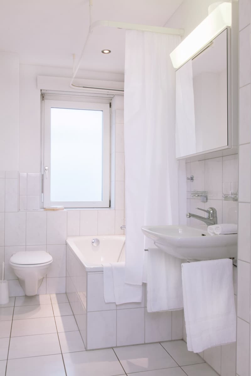 Bright and clean bathroom with white tiles, a toilet, a sink with a mirror cabinet, and a window providing natural light. A towel is hanging on the wall.