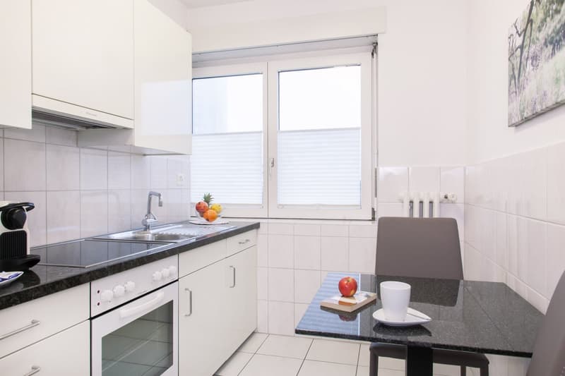Modern kitchen with white cabinets, granite countertop, built-in oven, and large windows providing natural light. The kitchen also features a dishwasher.