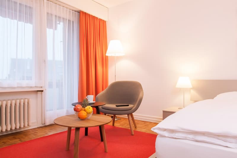 Spacious bedroom with large windows, orange curtains, wooden furniture including a chair and side tables, and a lamp. The room has a cozy and modern feel.