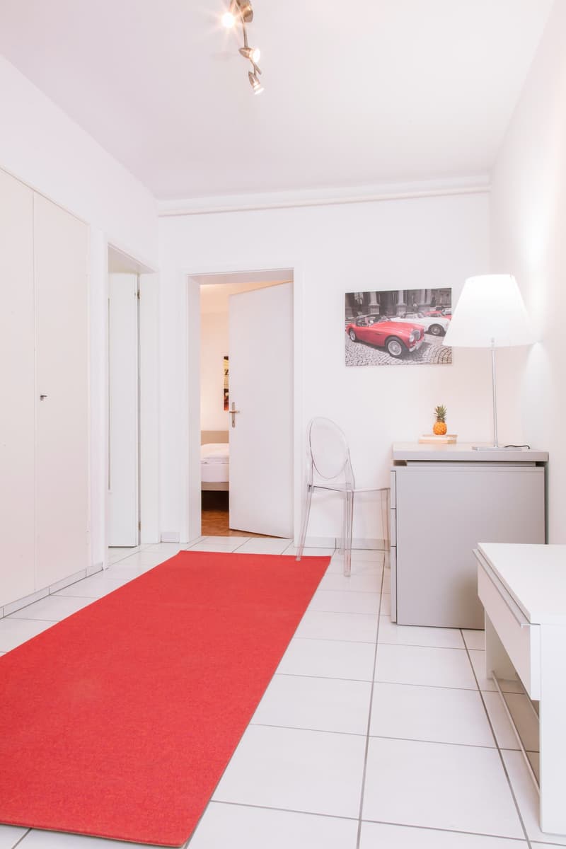 Bright and modern apartment with white walls, tile flooring, and a red carpet leading to the bedroom. The room features a desk with a lamp and a framed artwork of a red vintage car on the wall. The apartment appears to have a balcony or terrace, which is
