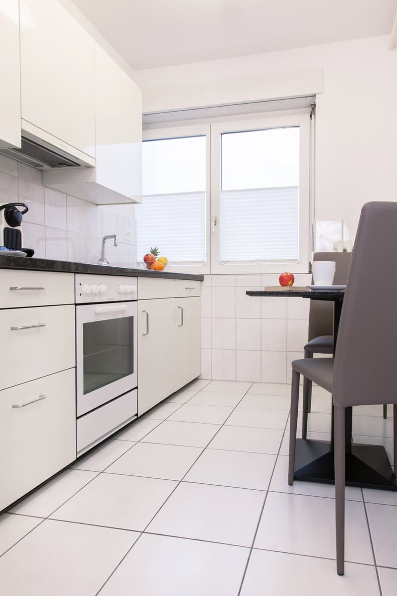 Modern kitchen with white cabinets, black countertops, tiled floor, and large windows. The kitchen includes a dishwasher and an oven.