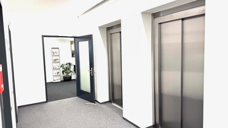 The image shows the interior of an office building lobby. There are two elevators visible, with a mirrored wall and a potted plant in the foreground. The floor is covered in a gray carpet, and the walls are white.