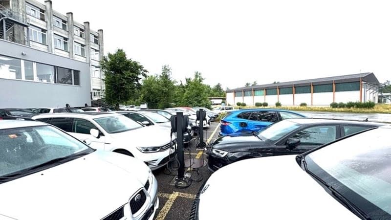 Outdoor parking lot with multiple cars parked, including a blue car and a white car. There is a building with multiple floors visible in the background.