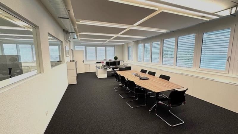 Large open office space with a conference table, chairs, and computer workstations. The room has large windows providing a view of the outside.