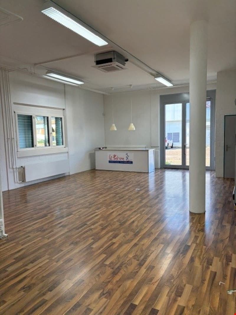 The image shows a large, open office space with a reception desk and hardwood flooring. The space has bright lighting from overhead fixtures and large windows, creating a modern and professional atmosphere.