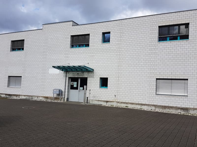A large, modern commercial or industrial building with a white brick exterior, multiple windows, and a covered entrance. The building has a flat roof and appears to be a single-story structure.