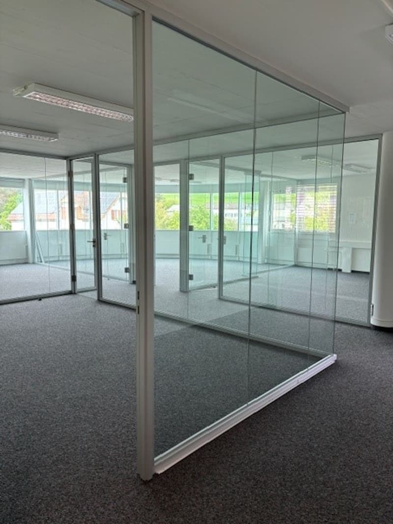The image shows a modern office space with glass walls and doors. The room has a carpeted floor and appears to be a large open-plan area with multiple glass partitions dividing the space. The glass walls allow natural light to flow through the office.
