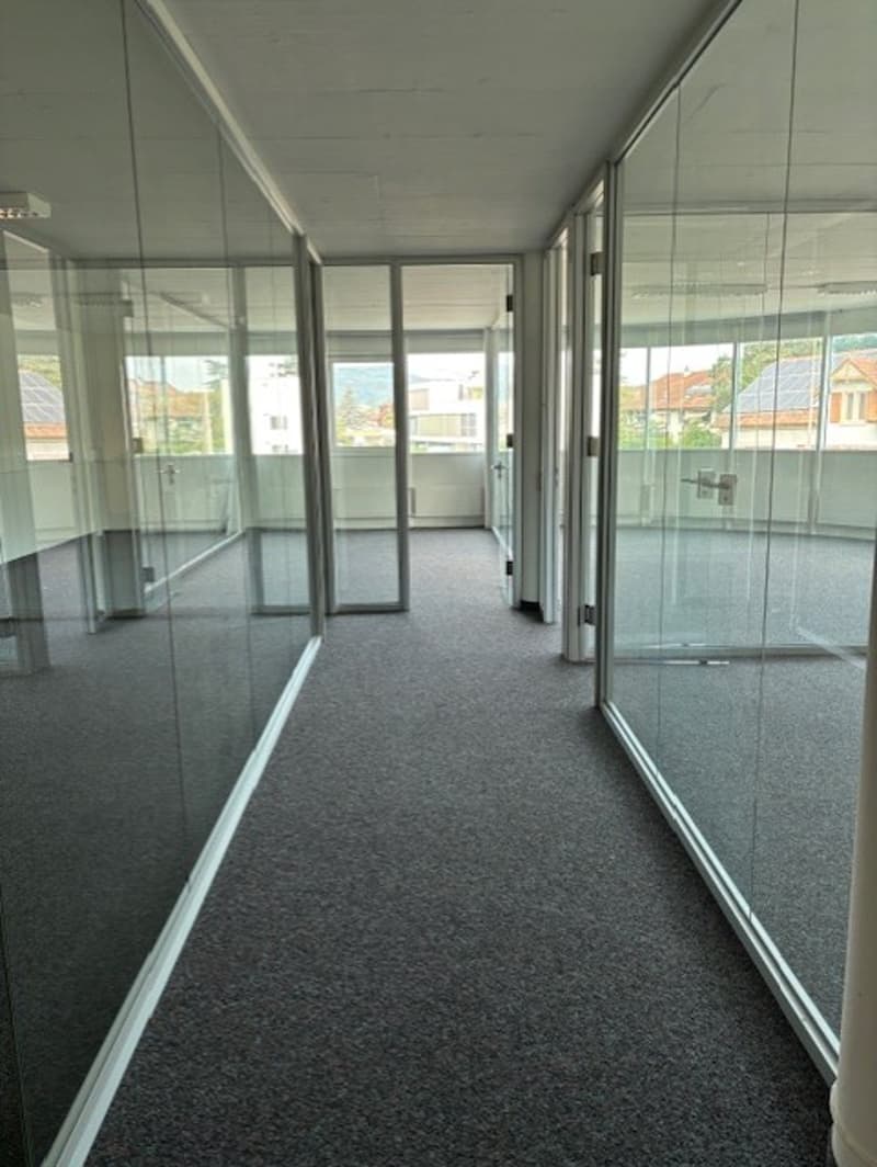 The image shows a long, narrow hallway or corridor with glass walls and doors on both sides. The floor is covered in a dark gray carpet. The space appears to be a commercial or office setting, with a clean and modern design.