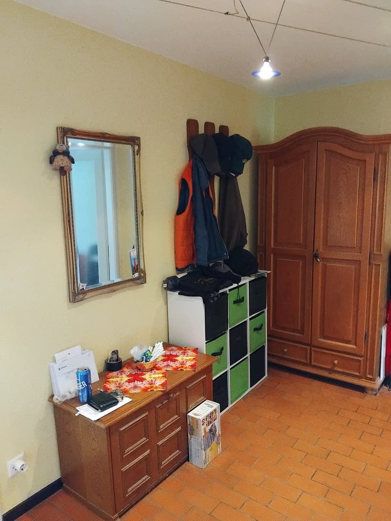 A room with a wooden cabinet, a mirror, a desk, and a storage box. The room has a tiled floor and a yellow wall. The wooden cabinet has a door and drawers. The mirror is mounted on the wall. The desk has a can, a wallet, and a box. The storage box is on t