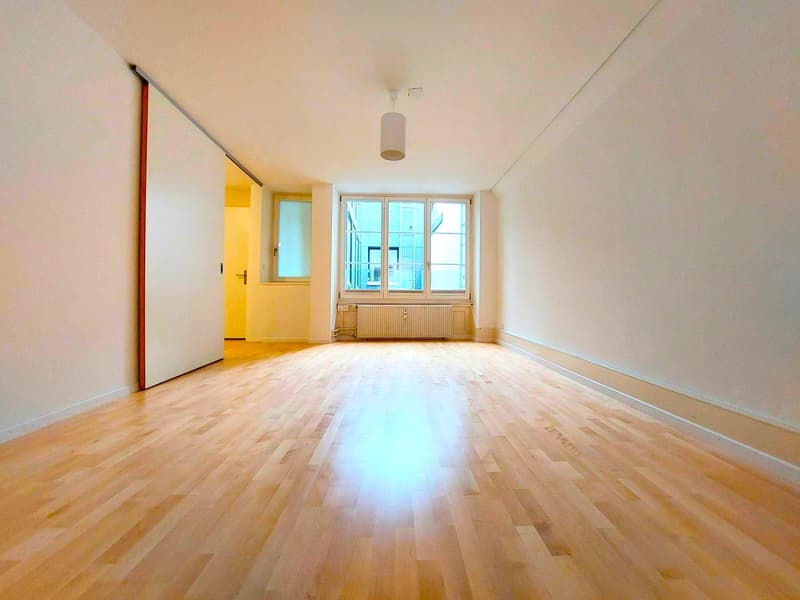 An empty room with wooden floors, white walls, a radiator, and large windows.