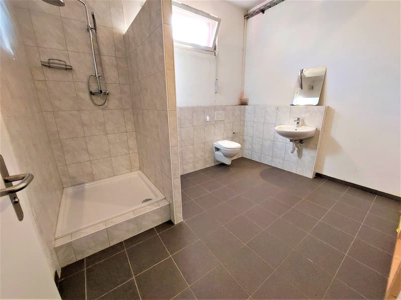 A modern and tidy bathroom featuring a shower, a toilet, and a sink against a tiled wall, complemented by natural light from a window and additional wall space.
