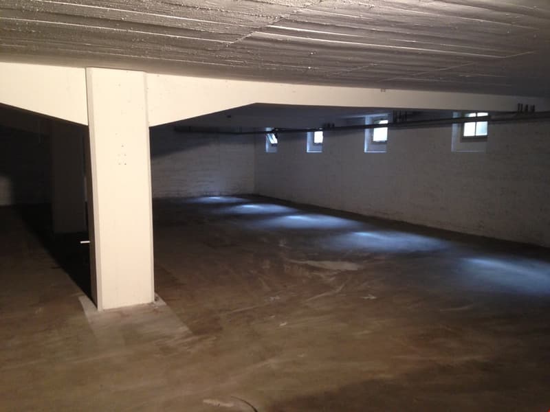 Empty parking lot, concrete floor, white walls, three small windows, central support pillar, overhead lighting.