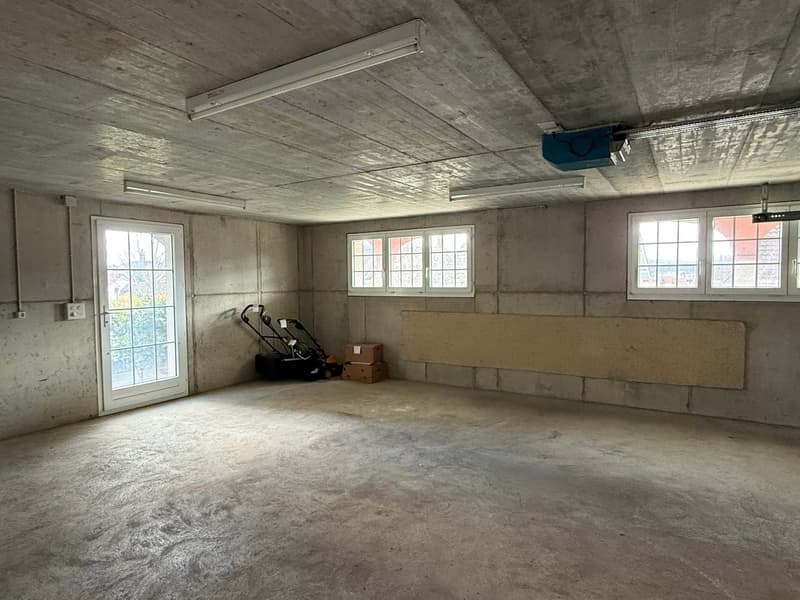 Empty room with concrete walls, ceiling, and floor, three windows, a door, and a ceiling-mounted light fixture, a lawn mower, and some boxes