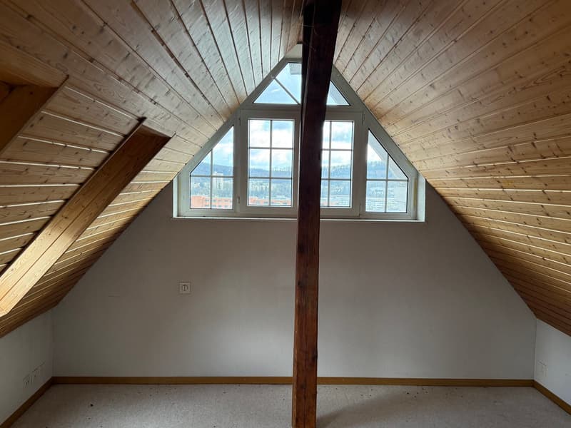 empty attic room with wooden ceiling, white walls, triangular window, power outlet, and a view of the city