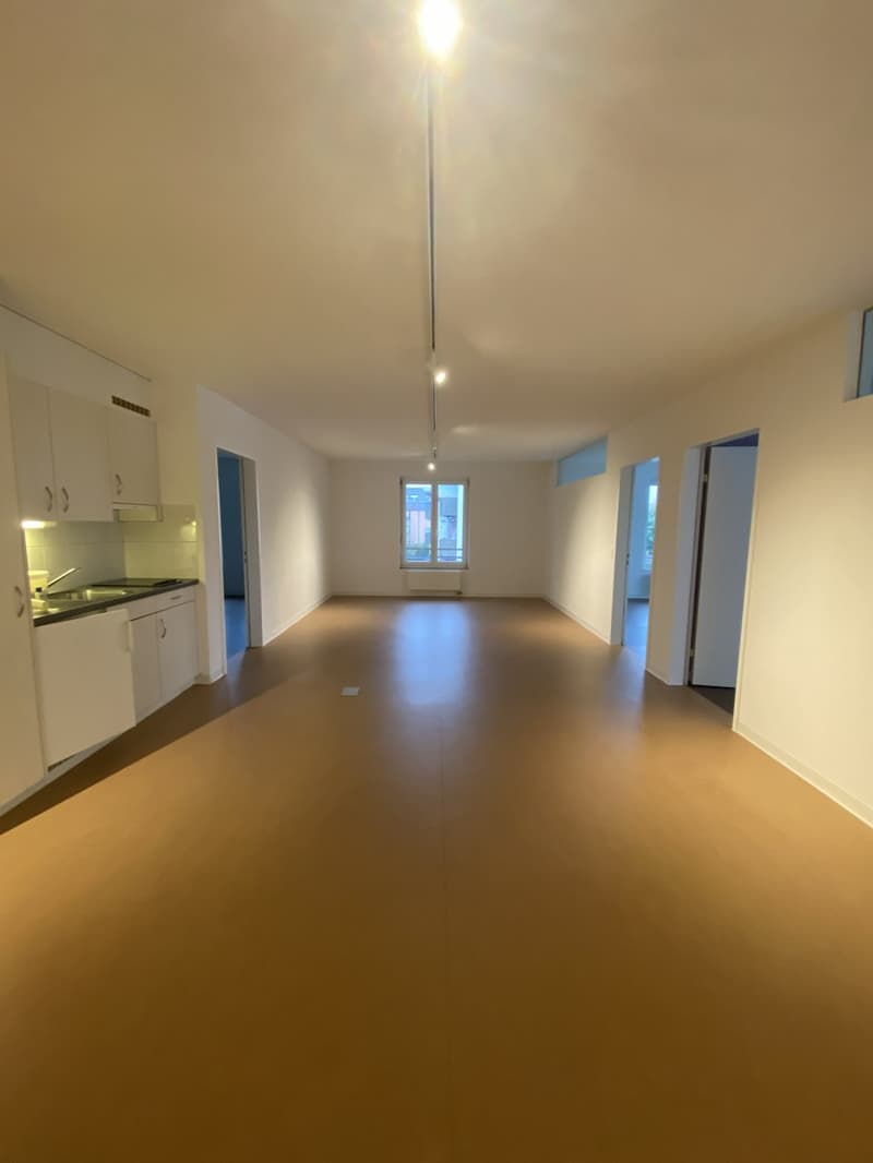 Empty room with white walls, brown floor, kitchen area with sink and cabinets, windows, and doors.