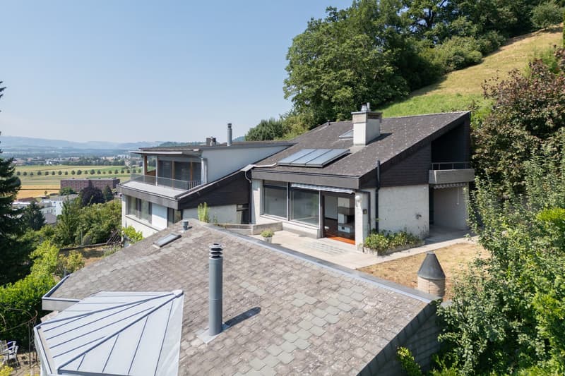 Modern house with solar panels, two levels, large windows, balcony, surrounded by trees and plants, view of the valley